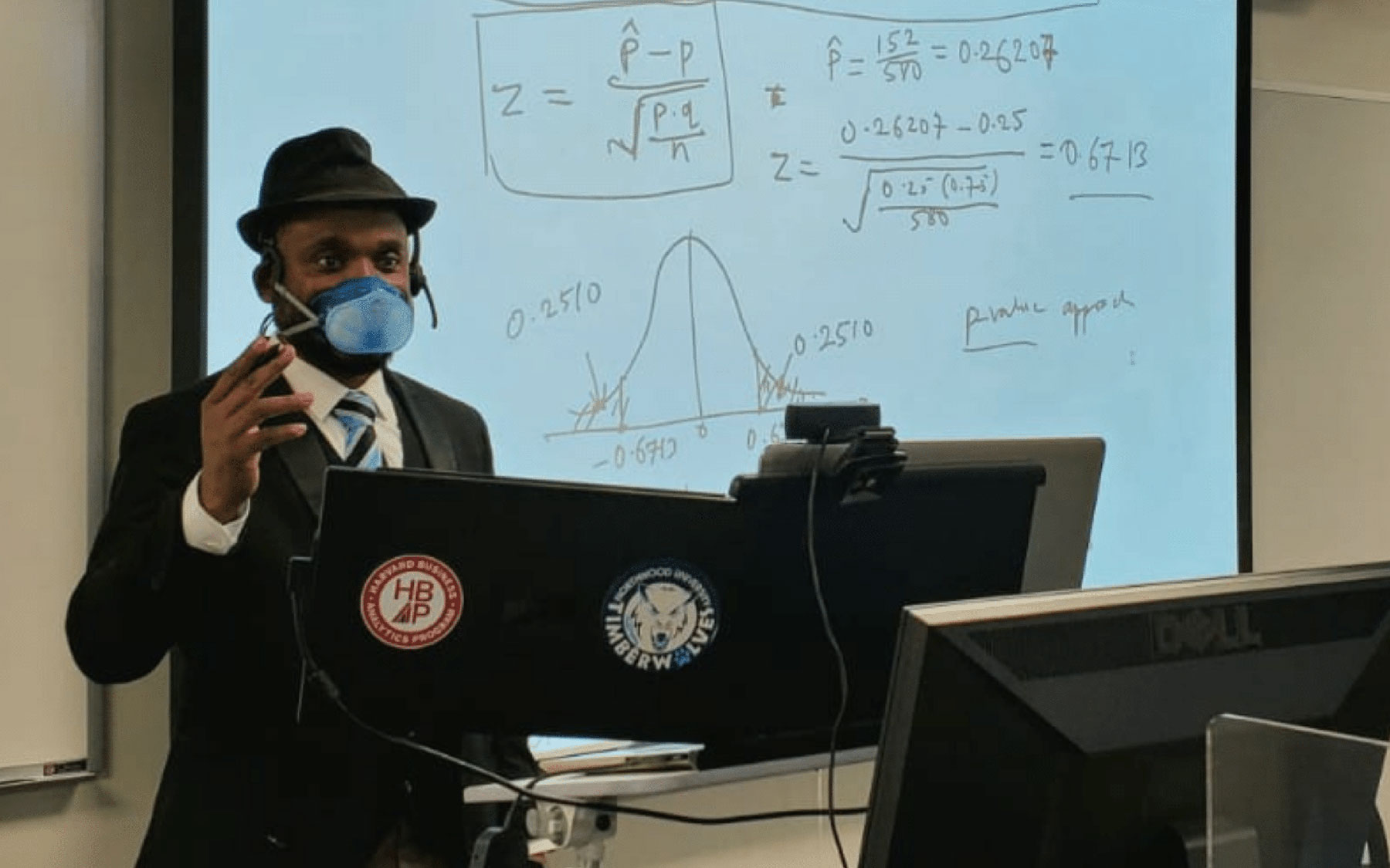 Dr. Itauma wears a mask and stands in front of a projector, teaching a class.